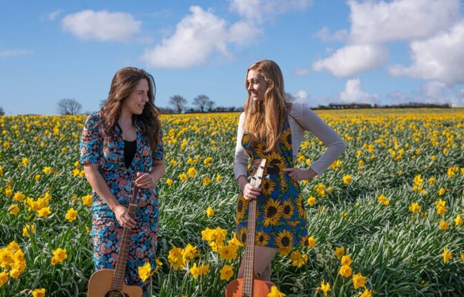 Two women holding guitars stood in a field of sunflowers